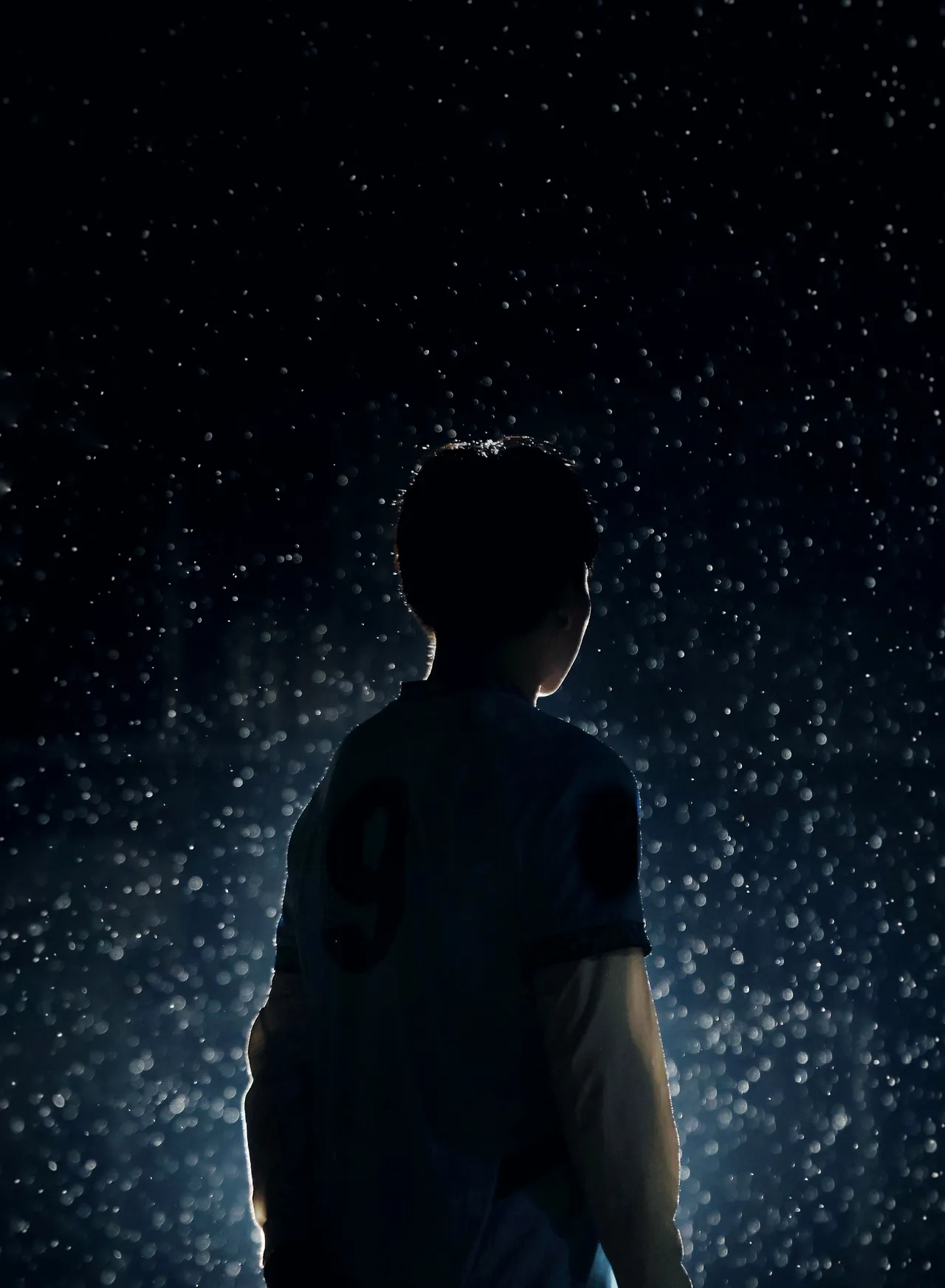 A footballer stands alone under a floodlight, rain falling through the beam.