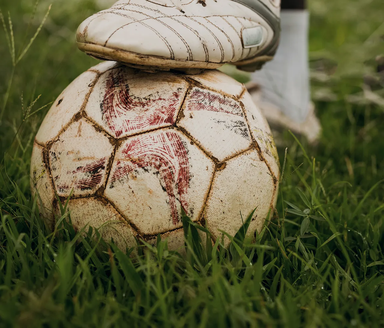 The toe of a white leather boot pinning a scuffed match ball to the grass.